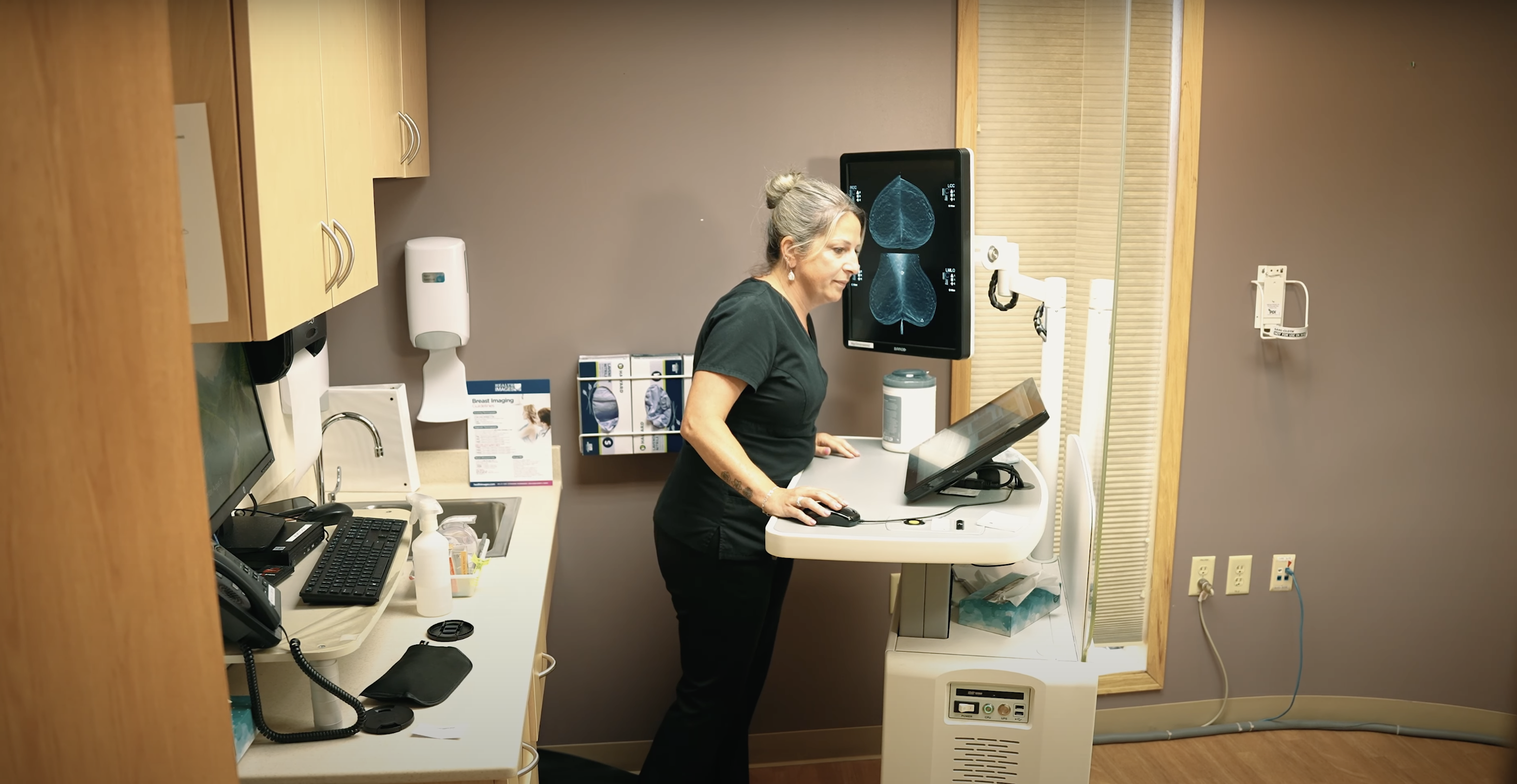 Breast cancer survivor and mammography technologist working in scrubs typing on computer with images of mammogram scans on the screen behind her.