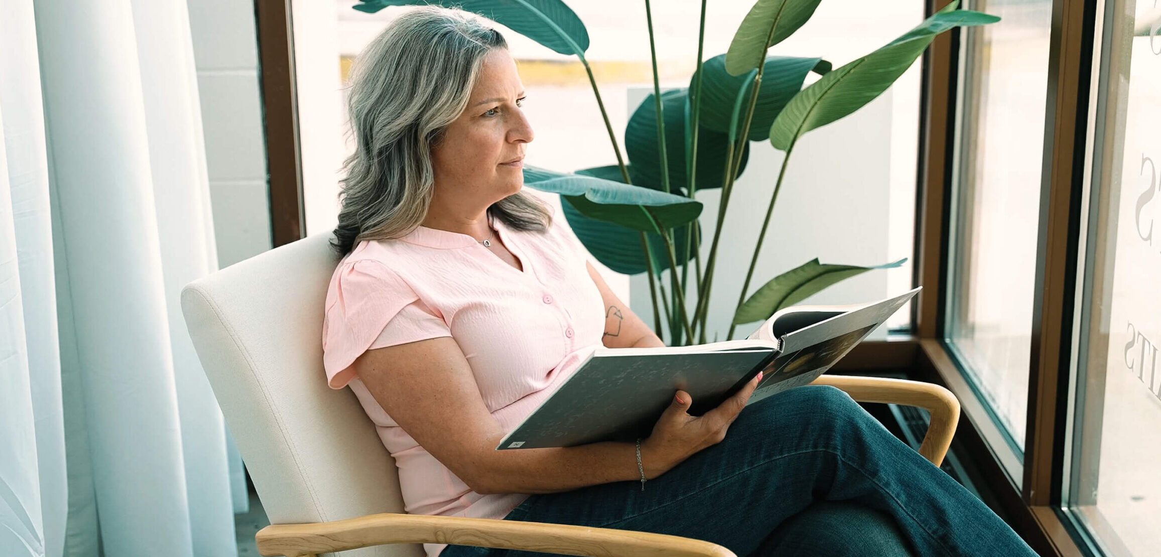 Kristin Miller, a breast cancer survivor and mammography tech at Envision Radiology sits by a window with a book in her lap.